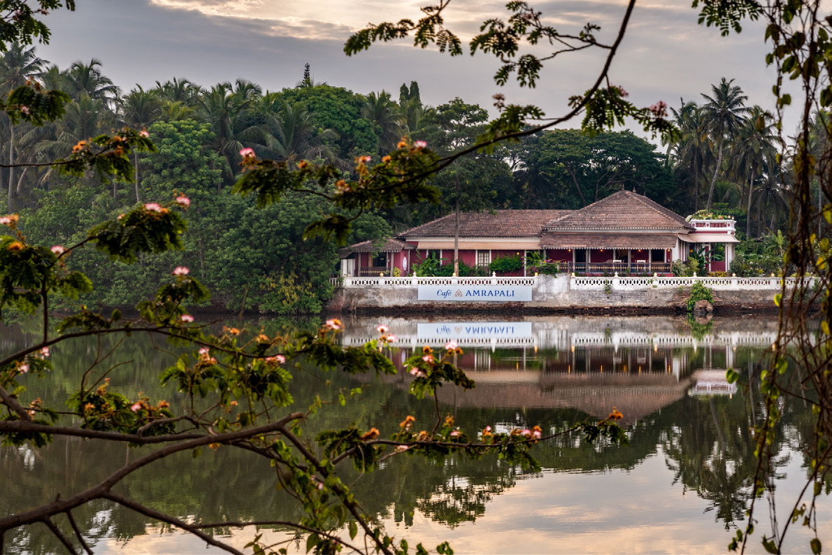 The house seen from across the water with its reflection and the Cafe Amrapali sign on the waterfront wall, framed by blossom. From the official e-brochure PDF, page 5.