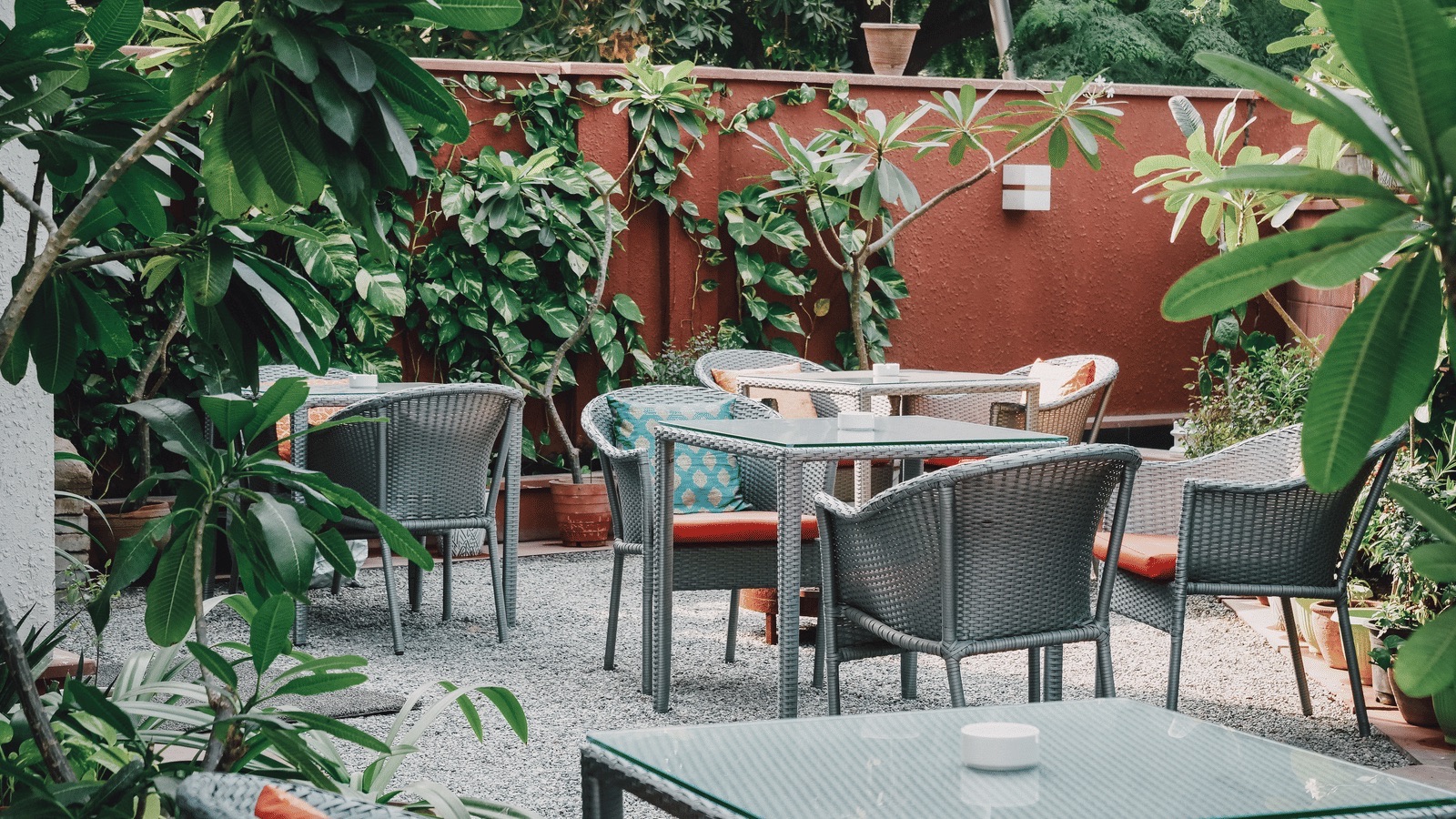 Courtyard alfresco dining area: rattan chairs and glass-top tables on gravel, red ochre wall covered in climbing plants, dense greenery