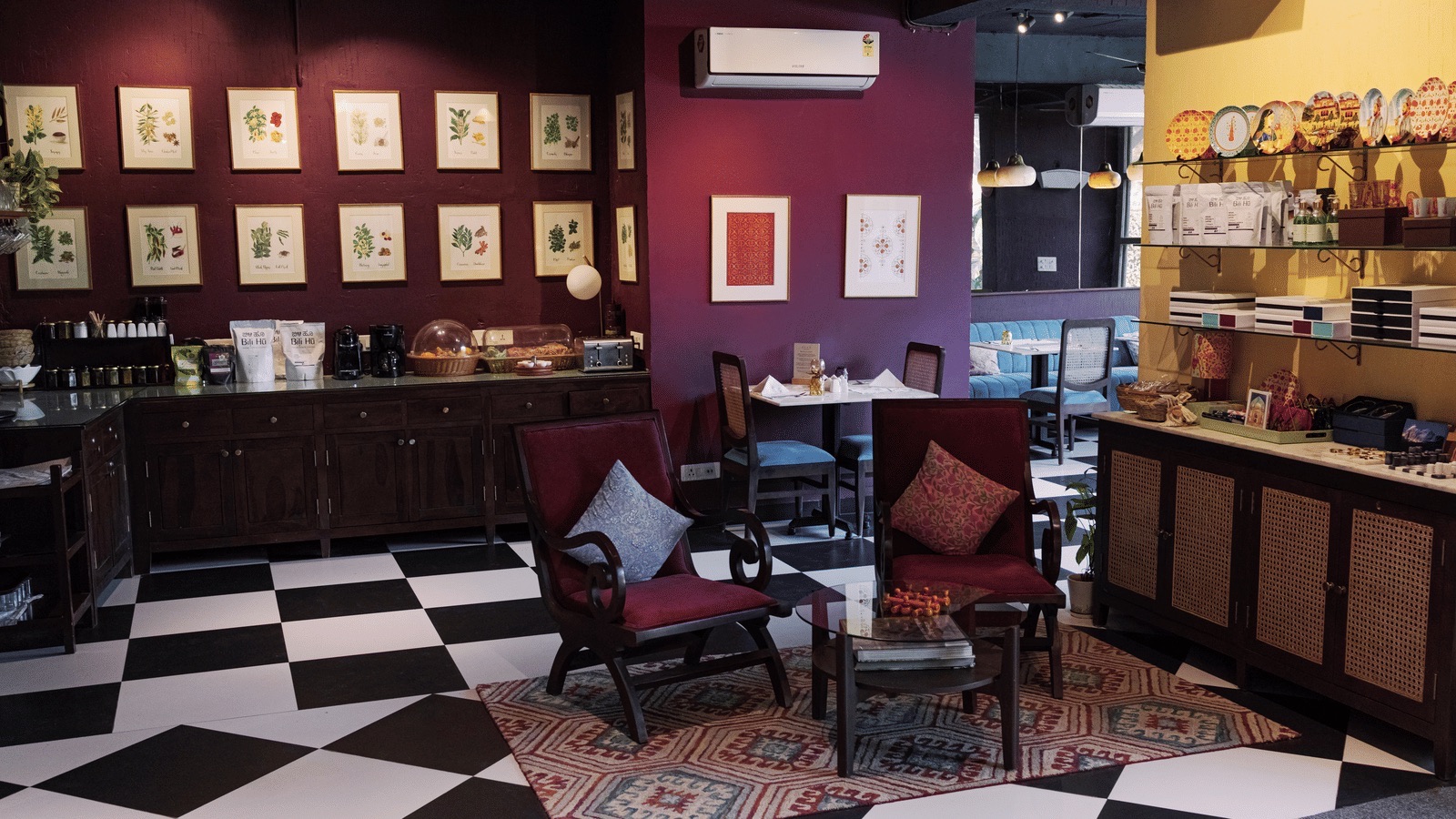 Indoor dining and lounge: aubergine walls with framed botanical prints, black-and-white chequerboard floor, kilim rug, wine-red armchairs, yellow retail shelf corner (Ekaa Boutique