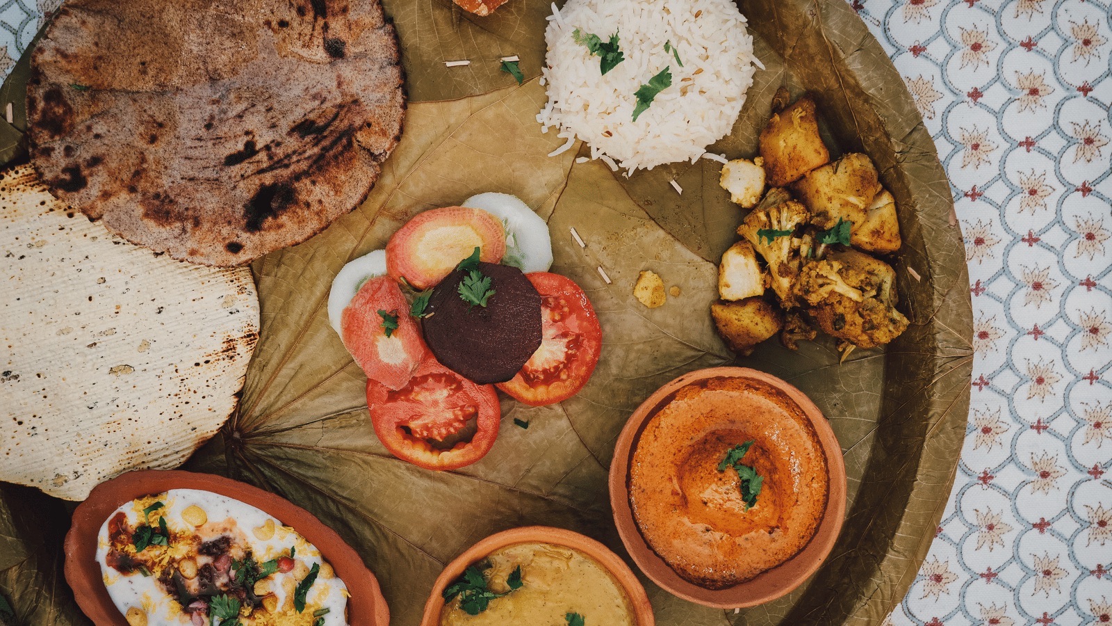 Overhead shot of a traditional thali served on a dried-leaf plate: rotis, rice, sabzi, dahi chaat, dal and curry in terracotta bowls, block-printed tablecloth